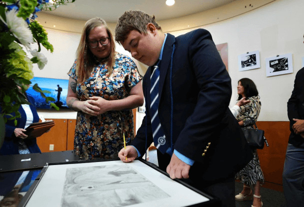 A young man in a school uniform signs a portrait under the watchful eye of a smiling woman in a floral dress.