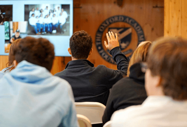 A person raises their hand in a classroom during a presentation, with other participants seated around and a projected image in the background.