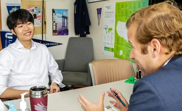 Two people are having a conversation in an office with a desk and chairs. One person is smiling, and various posters are on the wall.