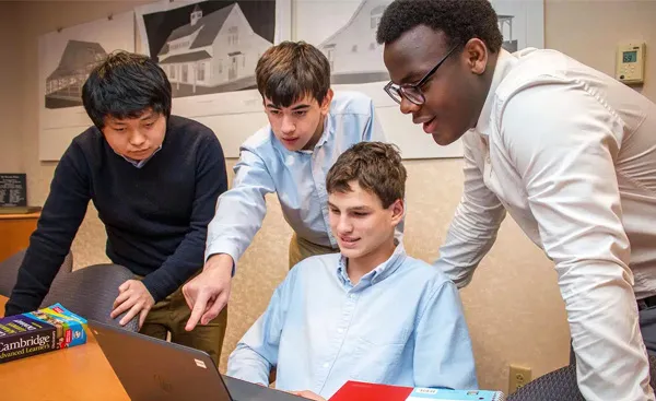 Four young men in a room look at a laptop, with one pointing at the screen. Books are on the table beside them. They appear to be collaborating on a project.