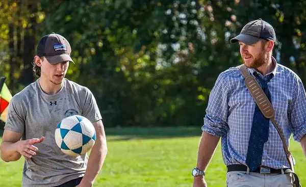 Two men walking outdoors; one in casual attire, dribbling a soccer ball, and the other in a shirt and tie, carrying a shoulder bag. Trees and grass are in the background.