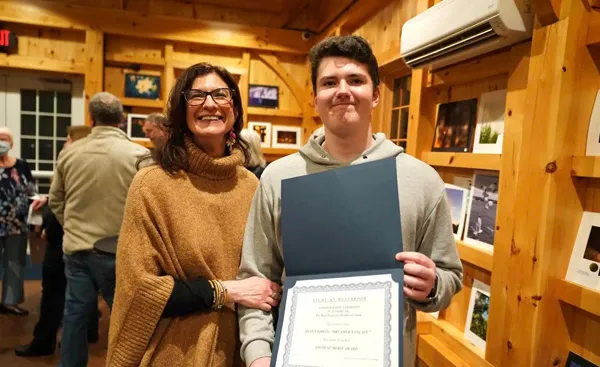 Two people in a wooden room. One holds an open certificate or diploma. Several people and framed photos are visible in the background.