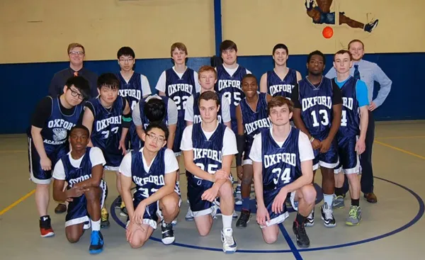 A basketball team poses in a gym, wearing matching "Oxford" jerseys. There are fourteen players and two coaches, with a basketball visible in the background.